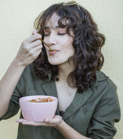 Wearing a green shirt, a curly-haired individual savors food from a pink bowl with eyes closed and contentment, embodying the essence of Savor & Nourish: A Mindful Eating Journey, set against a light yellow background.